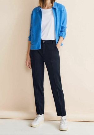 Lichtblauwe fleecejas over een wit T-shirt, gecombineerd met taps toelopende zwarte broek en witte sneakers, tegen een beige achtergrond.