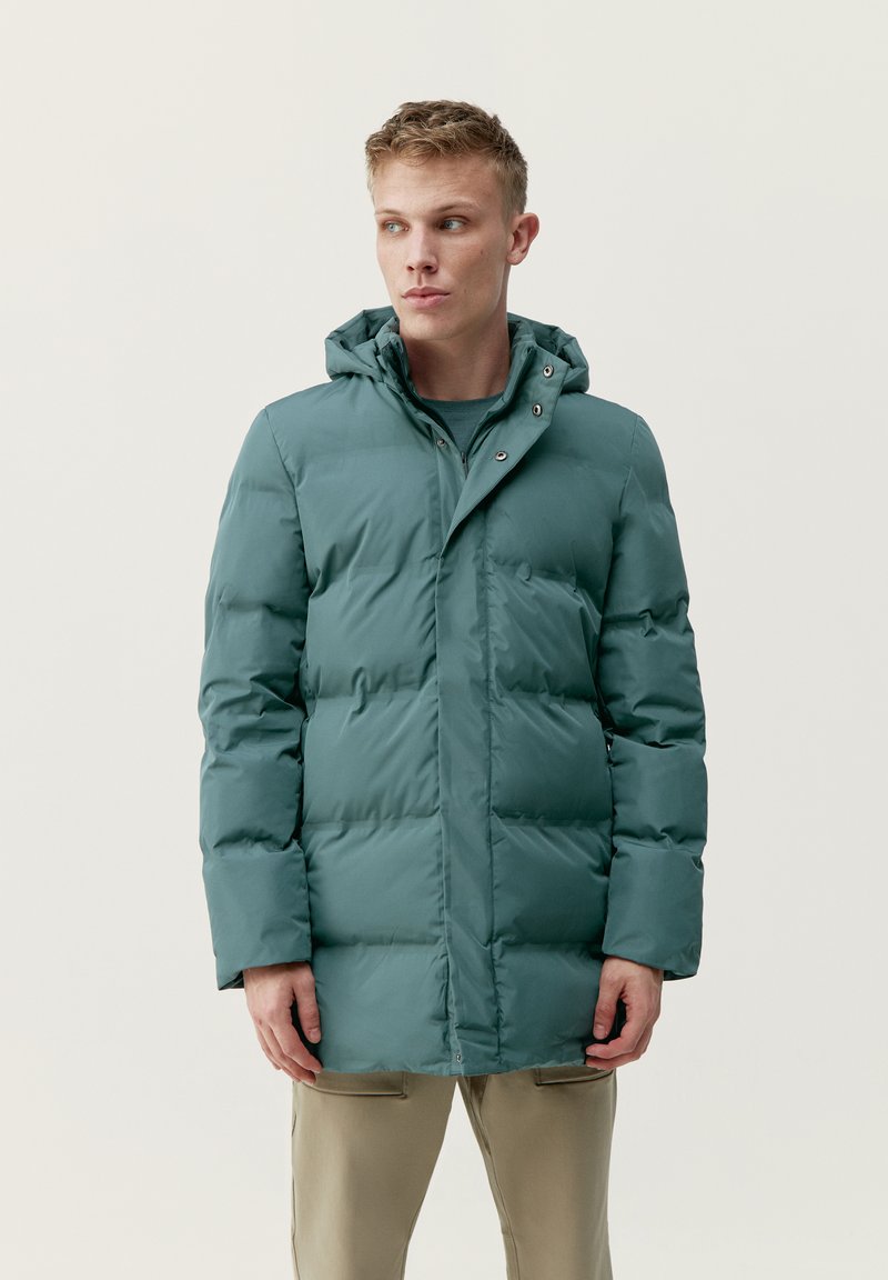 Padded jacket v tlmavo teal farbe, s kapucňou, predným zipsom, patentnými gombíkmi a hrubou prešívanou textúrou, spojená s béžovými nohavicami.