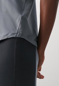 Primer plano de la cintura lateral y el brazo de una persona que lleva una camiseta gris y leggings oscuros, de pie contra un fondo claro y uniforme.