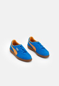 Modré semišové tenisky s oranžovými akcenty, hnědou gumovou podrážkou a šněrovacím designem. Logo PUMA na boku. Hladký povrch.