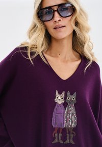 Fioletowy sweter z dzianiny z dekoltem w serek, ozdobiony haftem przedstawiającym dwa koty w płaszczach. Wyróżniające się detale obejmują wzory i fakturę.