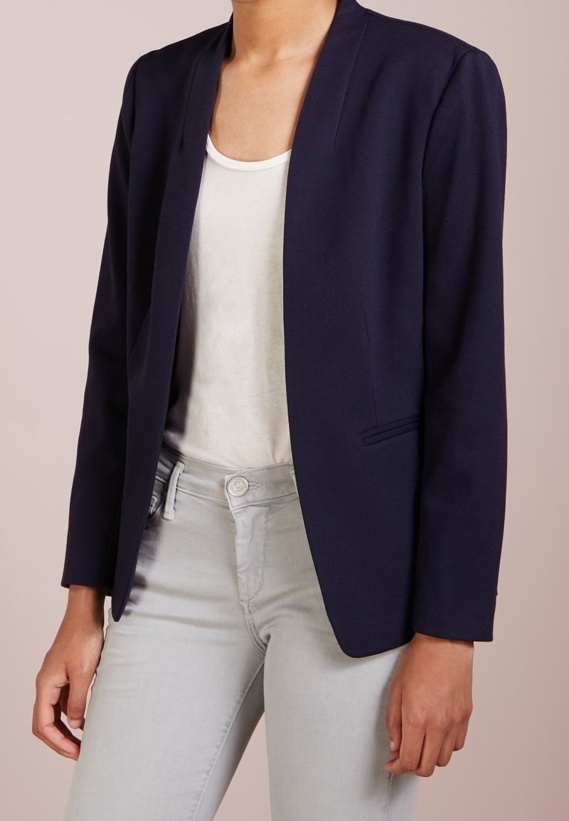 Femme portant un blazer bleu marine sur un haut blanc et un pantalon gris clair, debout devant un fond neutre.