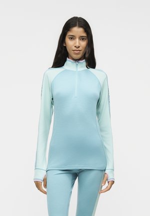Mujer que lleva una parte superior de manga larga con cremallera en azul claro y pantalones a juego, de pie frente a un fondo liso y claro.
