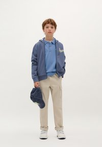 Modrá mikina na zip, světle modrá polo tričko, béžové cargo kalhoty, držící tmavě modrou čepici. Bílé tenisky s šedými akcenty. Jednoduchý design.