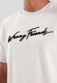 Camiseta de algodón blanca con un logotipo bordado en negro que dice "Wrong Friends" en el frente, escote redondo y mangas cortas.