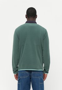 Groene lange mouwen blouse met een donkere kraag en lichtblauwe afwerking aan de zoom. Zacht materiaal en relaxte pasvorm, met een naaddetail aan de achterkant.