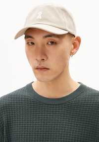 Casquette de baseball en coton beige clair avec une visière incurvée et un logo brodé blanc. Portée par une personne portant un pull texturé vert foncé.