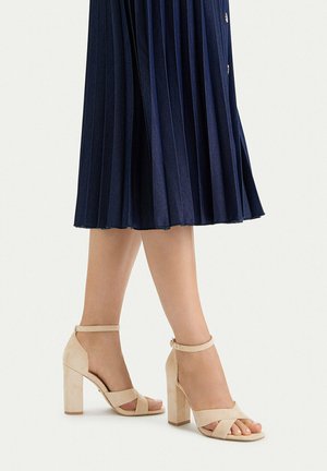 Piernas con sandalias de tacón altas beige con correas en el tobillo combinadas con una falda midi plisada azul marino sobre un fondo blanco.