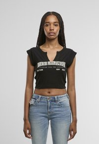 Top corto negro de tirantes con escote en V, textura acanalada y texto en blanco "True Religion". Combinado con jeans ajustados de corte slim en azul claro.
