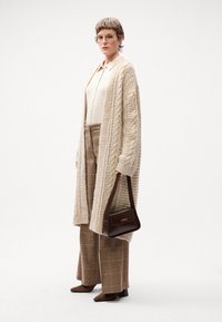Cardigan lavorato a maglia beige, blouse crema, pantaloni a quadri marroni, borsa marrone e décolleté marroni con punta. Focus sulla texture e sui toni neutri.