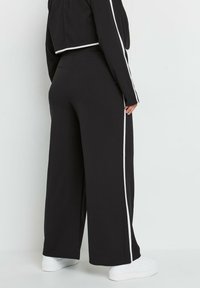 Czarne spodnie szerokie z białymi paskami po bokach, wykonane z miękkiego materiału. Ich design ma wysoki stan i luźny krój, idealnie komponując się z białymi trampkami.