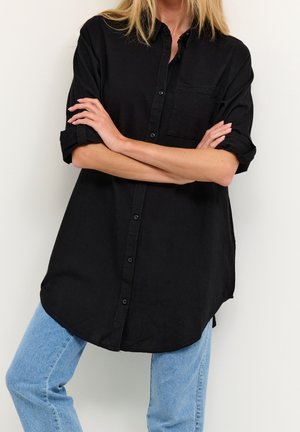 La personne porte une chemise noire à manches longues boutonnée, les manches retroussées jusqu'aux coudes, ainsi qu'un jean bleu clair, les bras croisés sur la poitrine contre un mur blanc.