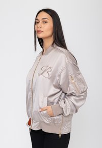 Vrouw met lang donker haar die een lichtbeige satijnen bomberjack draagt met een "B"-logo en ritszakken, handen in de voorzakken, zijwaarts kijkend.