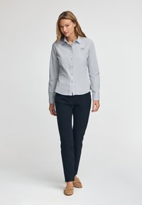 Ljust grå button-down skjorta med långa ärmar, kombinerad med mörk marinblå byxor. Har en liten logotyp Patch på skjortan. Slät textur.