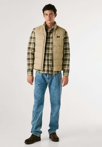 Gilet imbottito beige con colletto alto sopra una camicia a quadri verde e blu, abbinato a jeans azzurri chiari e stivaletti marroni.