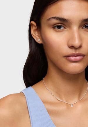 Collana in argento con ciondolo a forma di piccolo orso, abbinata a orecchini a perno: una perla bianca e un piccolo orso, indossata con un top di colore azzurro chiaro.