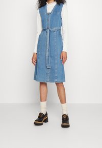 Denimmekko, jossa on tiukka yläosa, A-linjainen hame ja säädettävä vyö. Ominaisuuksiin kuuluvat napitettu etuosa, sivutaskut ja vaaleansininen väri. Mustat platform-kengät.