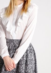 Blouse blanche avec un devant froncé, des manches longues et une fermeture à boutons. Associée à une jupe gris foncé avec un motif floral.