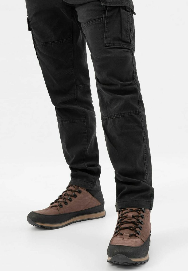 Persona con pantaloni cargo neri e scarpe da trekking marroni con suole nere e lacci a fantasia, in piedi su uno sfondo bianco.