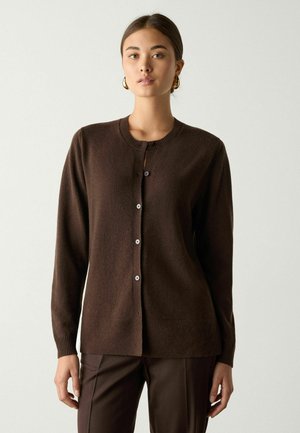 Femme portant un cardigan boutonné marron foncé et un pantalon assorti, debout devant un fond clair uni, cheveux tirés en arrière, boucles d'oreilles créoles dorées.