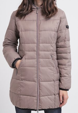 Femme portant une longue veste matelassée mauve avec une fermeture éclair devant et deux poches latérales zippées, les mains partiellement dans les poches.