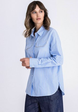 Vrouw met golvend haar, gekleed in een lichtblauw-wit verticale gestreept overhemd met knopen en twee borstzakken, en een donkere broek, die zijwaarts kijkt.