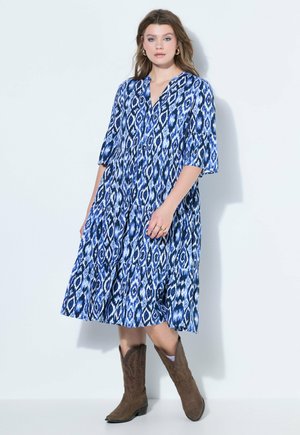 Kobieta w niebiesko-białą, wzorzystą sukienkę do połowy łydki z rękawami do łokcia oraz brązowe, haftowane kowbojskie buty, stojąca przy białej ścianie.