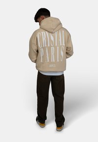 Beige hoodie met grote witte tekst "CRYSTAL PARIS" op de achterkant, gedragen over een wit shirt, gecombineerd met donkere broek en sneakers.