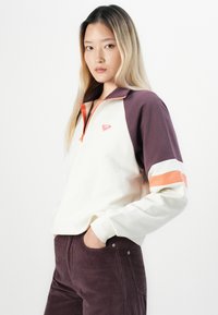 Jonge vrouw met lang blond haar, gekleed in een wit en paarse zip-up sweatshirt met oranje accenten en paarse corduroy broek, met handen in haar zakken.