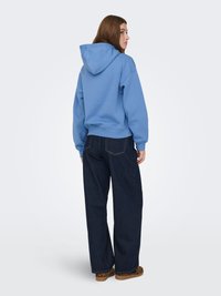 Blauwe hoodie met capuchon, losse pasvorm, van katoen; gecombineerd met wijde donkerblauwe spijkerbroek en pantoffels met luipaardprint. Gezien vanaf de achterkant.