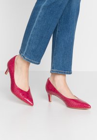 Talons en cuir verni rose vif avec une texture crocodile, bout pointu et petit talon, associés à un jean cropped en denim bleu.