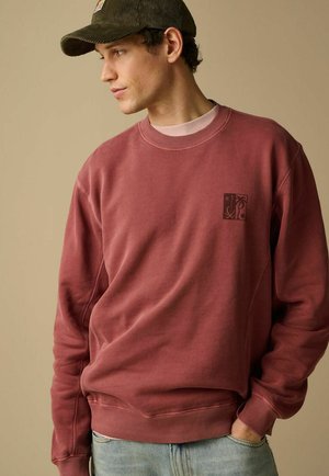 Jonge man die een bordeauxrode sweater draagt met een kleine zwarte afbeelding op de borst en een donkergroene corduroy pet, kijkend naar links.