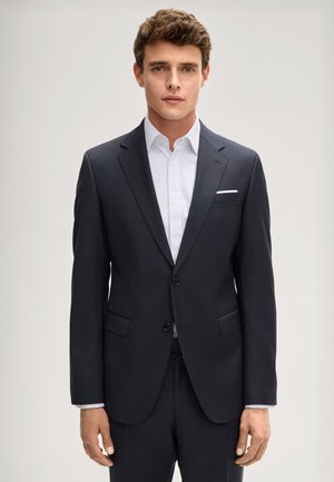 Młody mężczyzna w ciemnym, szytym na miarę garniturze i białej koszuli, stojący twarzą do przodu na jednolitym jasnym tle.