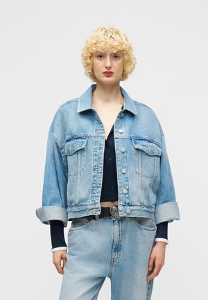 Νεαρός με κοντά σγουρά ξανθά μαλλιά που φοράει ανοιχτό μπλε τζιν μπουφάν και τζιν παντελόνι με ψηλή μέση, σε απλό φόντο.