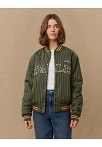 Grüne gestreifte Bomberjacke mit weißem "YALE"-Schriftzug, gerippten orangen Akzenten und Druckknöpfen. Getragen über einem weißen Hemd und blauen Jeans.
