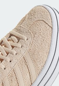 Beige sneaker with textured suede upper, beige laces, three decorative stripes, and a white rubber sole accentuated by zigzag stitching.