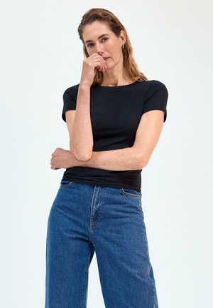 Kvinde med lys hud og vådt hår iført en sort tætsiddende kortærmet top og højtaljede blå jeans, poserer med den ene hånd nær munden.