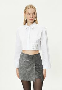 Weißes, kurzes Hemd mit langen Ärmeln, Knopfleiste und einem mit Schmuck akzentuierten linken Teil. Kombiniert mit einem silbernen Glitzerwickelrock.