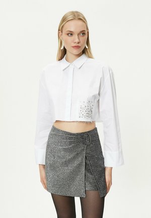 Camisa blanca corta con mangas largas, cierre de botones en la parte delantera y adorno de joyas en el lado izquierdo. Combinada con una falda corta de purpurina plateada.