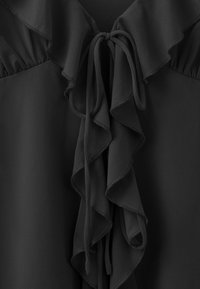 Blouse noire avec devant à volants et fermeture par lien au niveau du col, confectionnée dans un tissu léger avec des détails froncés aux épaules.