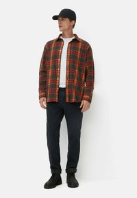 Rood en zwart geruit flanellen shirt over een wit T-shirt, gecombineerd met donkere broeken en zwarte schoenen. Model met een zwarte pet, staande pose.