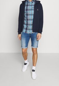 Marinblå zip-up-hoodie över en rutmönstrad skjorta med blå, gröna och vita mönster, kombinerad med uppvikta jeansshorts och vita sneakers.