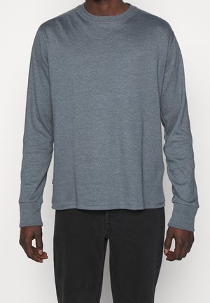 Man wearing a plain, long-sleeve gray crewneck shirt and black pants against a white background.