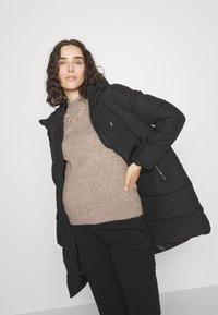 Chaqueta de plumas negra con capucha, que presenta un cierre frontal, puños elásticos y bolsillos laterales, llevada sobre un suéter beige.