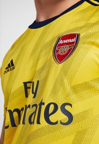 Yellow Arsenal football jersey with club crest, Adidas logo, and Fly Emirates sponsor branding on a person’s upper torso.