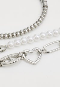 Three bracelets: one silver beaded, one white pearl, and a silver chain featuring a heart-shaped link. Smooth textures, shiny finish.