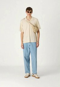 Ung mann iført beige polo skjorte, lys blå jeans, hvite joggesko, solbriller, og beige crossover-veske som står mot en ensfarget bakgrunn.