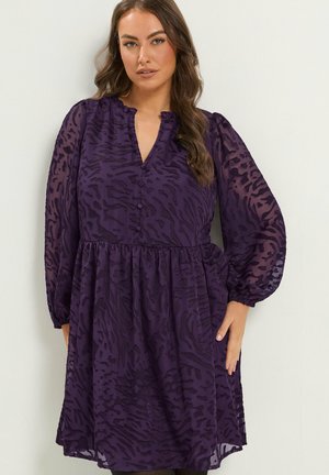 Femme aux longs cheveux bruns portant une robe violette à manches longues et motif, avec des boutons et une taille froncée, debout devant un fond clair.