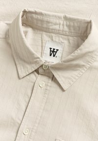 Beige overhemd met kraag en een gestructureerd gestreept patroon, voorzien van witte knopen en een label met een zwart logo. Glad stof met een subtiele glans.