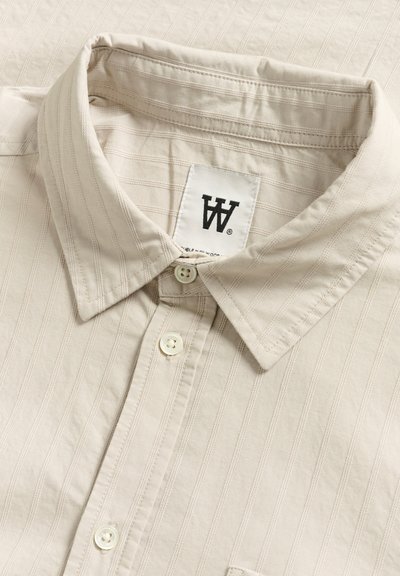 Chemise beige à col avec un motif rayé texturé, dotée de boutons blancs et d'une étiquette avec un logo noir. Tissu lisse avec un léger éclat.